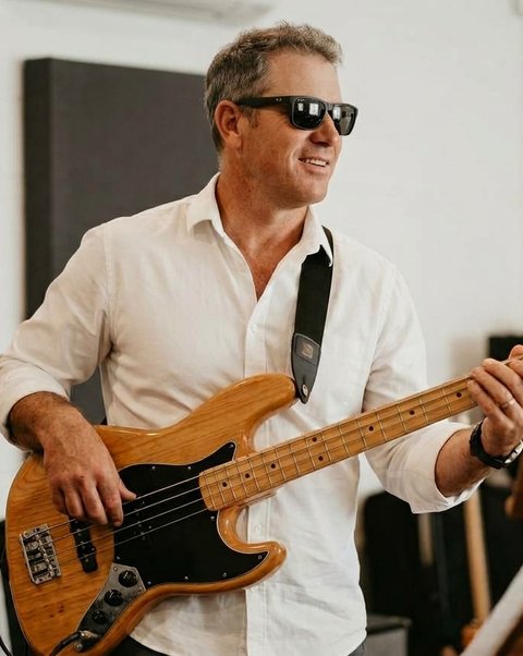Smiling across the room, holding a natural-finish bass.