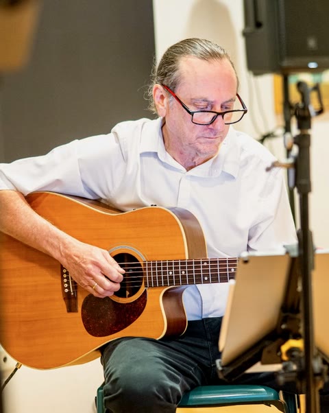 Seated with an acoustic guitar, reading off a music stand.