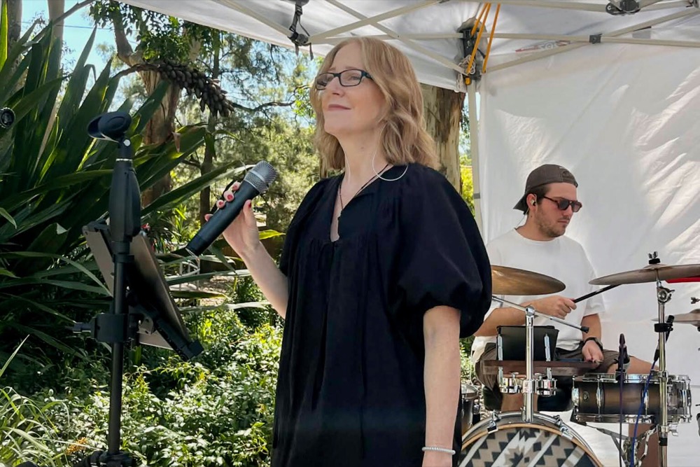 Emma singing into a handheld mic at an outdoor market, with Josh on drums under the marquee behind her.